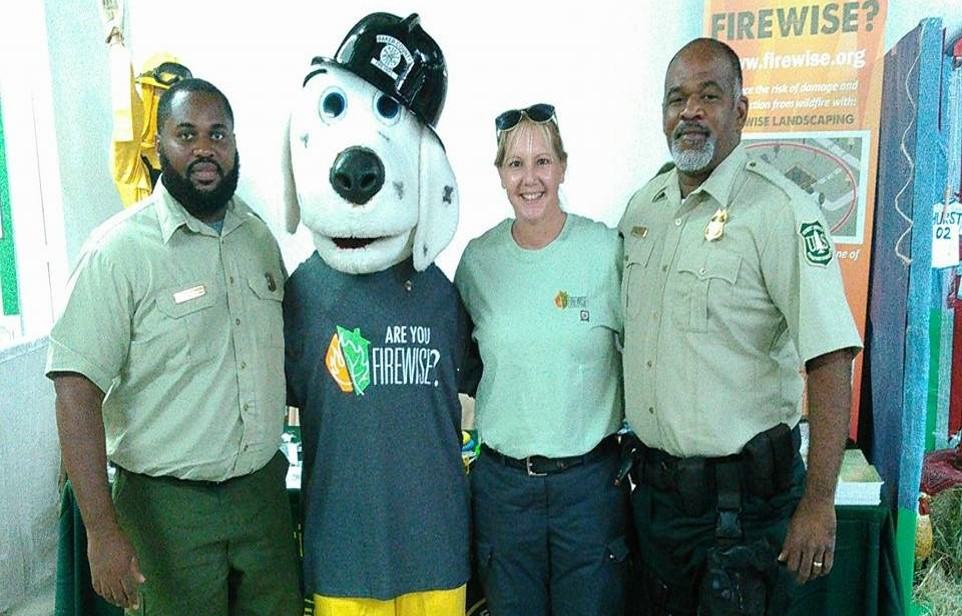 Firewise coordinator posing with Sparky mascot and two USFS staff.