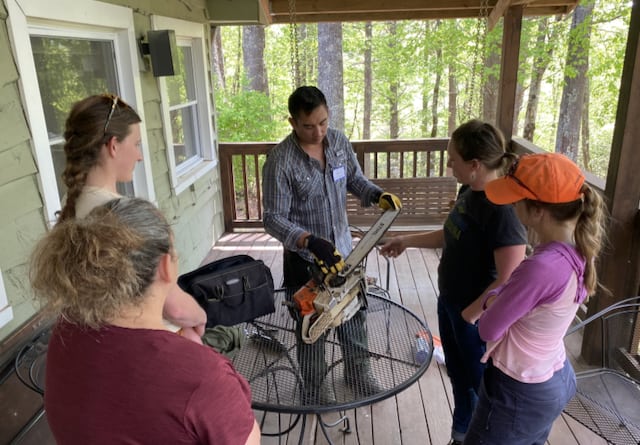 Five people stand around a table on a screened porch. One person facing the camera holds a chainsaw on the table and shows the group a particular feature of it.