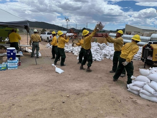 Eight people in firefighting gear stand in a line outdoors as they pass sandbags between each other to form a pile.
