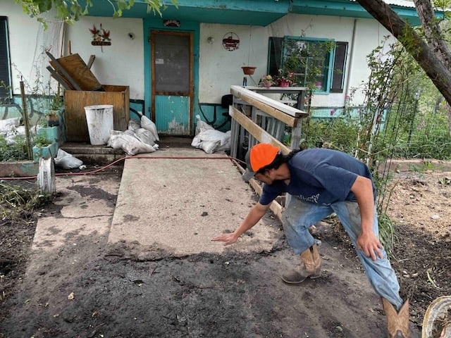 A man in workwear points to a broken cement walkway leading up to the front door of a flooded home. There is mud in the foreground and several bags of debris in front of the door.