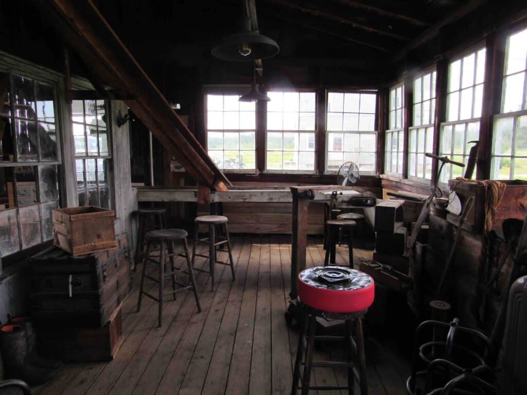 Interior photo of an old wooden barn, with glass windows looking outdoors, and a variety of stools and chairs in the room.